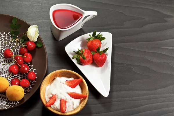 On a dark wooden background there is an original mug with tea, a decorative dish, a wooden cup, strawberries, sweet cherries, sweets, yogurt, cookies.