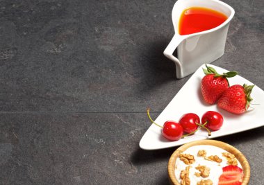 Koyu ahşap arka planda çay, dekoratif bir çanak, ahşap bir fincan, çilek, tatlı kiraz, tatlılar, yoğurt, kurabiye ile orijinal bir kupa var.