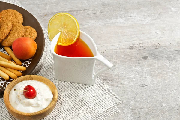 On a dark wooden background there is an original mug with tea, a decorative dish, a wooden cup, strawberries, sweet cherries, sweets, yogurt, cookies.