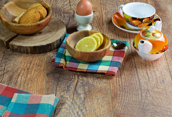 On a dark wooden background is an original mug with tea and a teapot, a decorative dish and a napkin, an egg on a stand, a wooden cup, sweets, cookies,