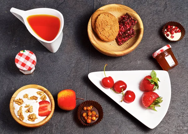 On a dark wooden background there is an original mug with tea, a decorative dish, a wooden cup, strawberries, sweet cherries, sweets, yogurt, cookies.