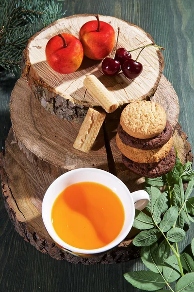 On a light wooden table there is an original mug with a cup of tea, a  a wooden cup, a wooden stand, a sweet cherry, sweets, and cookies.