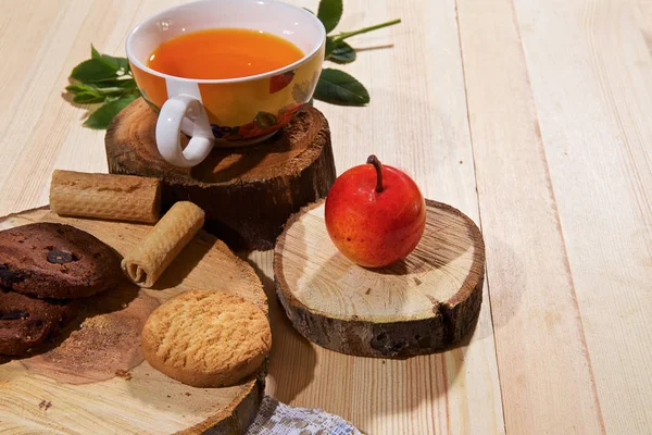 On a light wooden table there is an original mug with a cup of tea, a teapot, a wooden cup, a wooden stand, a sweet cherry, sweets, and cookies.