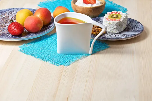 On a light wooden background - an original mug with tea, a decorative dish, a wooden cup, various fruits, cake, yogurt, cookies.