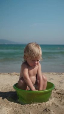 Mutlu çocuk plajda yaz tatilinin tadını çıkarıyor. Kıvırcık saçlı çocuk kumsaldaki yeşil havuzda mutlu bir şekilde oynuyor. Küçük neşeli çocuk güneşli gökyüzünün altında yumuşak deniz dalgalarıyla
