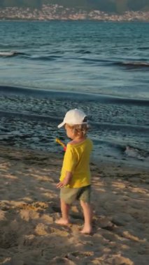 Parlak sarı tişörtlü ve beyaz şapkalı küçük bir çocuk gün batımında sahilde yürüyor, sakin sahil manzarasının ve anın keyfini çıkarıyor.