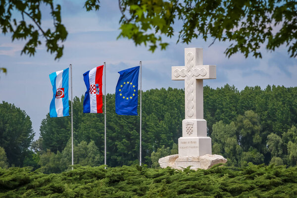 Вид на крестовый памятник, который говорит жертвам войны за свободную Хорватию в память о хорватских защитниках войны с поднятыми рядом с ним флагами в Вуковаре, Хорватия
.