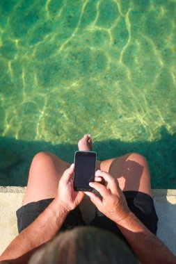 Sahil iskelede otururken Smartphone tutan bir adam.