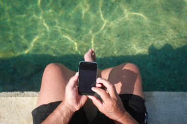 Sahil iskelede otururken Smartphone tutan bir adam.