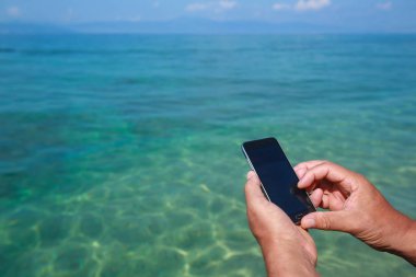 Sahil iskelede otururken Smartphone tutan bir adam.