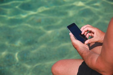 Sahil iskelede otururken Smartphone tutan bir adam.