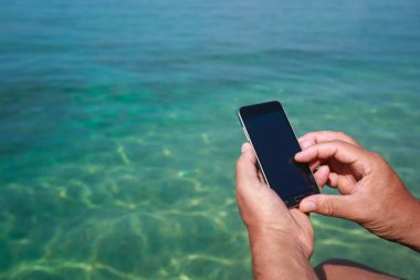 Sahil iskelede otururken Smartphone tutan bir adam.