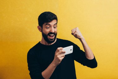 Sarı arka plan üzerinde izole cep telefonunda oyun oynamayı neşeli bir genç adam portresi kutluyor