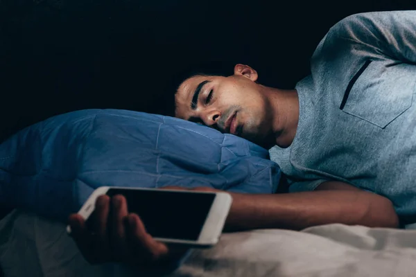 Um homem a dormir na cama e a segurar um telemóvel. Foto de conceito de vício em telefone inteligente . — Fotografia de Stock