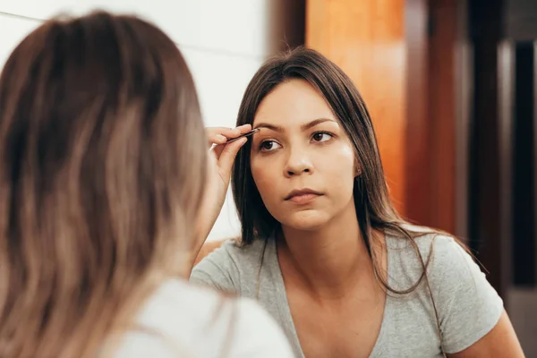 若い女性が鏡の前でピンセットで眉毛を摘採 — ストック写真