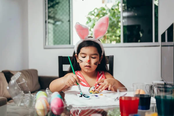Счастливой Пасхи! Красивая девочка рисует пасхальные яйца. Счастливая семья готовится к Пасхе. Милая маленькая девочка с кроличьими ушами на Пасху — стоковое фото