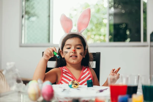 Счастливой Пасхи! Красивая девочка рисует пасхальные яйца. Счастливая семья готовится к Пасхе. Милая маленькая девочка с кроличьими ушами на Пасху — стоковое фото