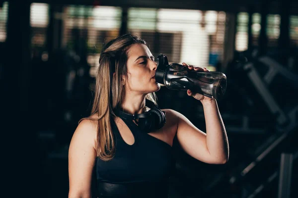 Una mujer joven tomando un descanso en el gimnasio con una botella de agua o bebida energética —  Fotos de Stock