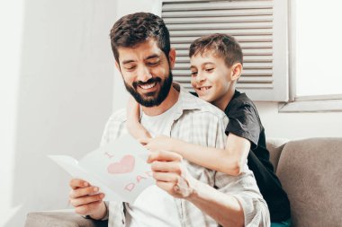 Babalar günün kutlu olsun! Oğlum babamı tebrik ediyor ve ona tebrik kartı veriyor. Baba ve oğul gülümsüyor ve sarılıyorlar. Aile tatili ve birliktelik.