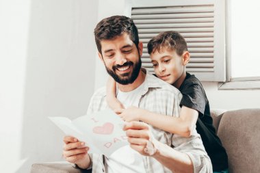 Babalar günün kutlu olsun! Oğlum babamı tebrik ediyor ve ona tebrik kartı veriyor. Baba ve oğul gülümsüyor ve sarılıyorlar. Aile tatili ve birliktelik.