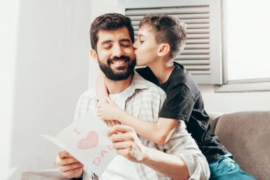 Babalar günün kutlu olsun! Oğlum babamı tebrik ediyor ve ona tebrik kartı veriyor. Baba ve oğul gülümsüyor ve sarılıyorlar. Aile tatili ve birliktelik.