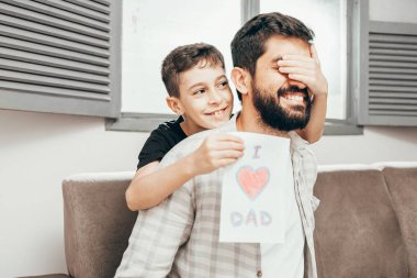 Babalar günün kutlu olsun! Oğlum babamı tebrik ediyor ve ona tebrik kartı veriyor. Baba ve oğul gülümsüyor ve sarılıyorlar. Aile tatili ve birliktelik.
