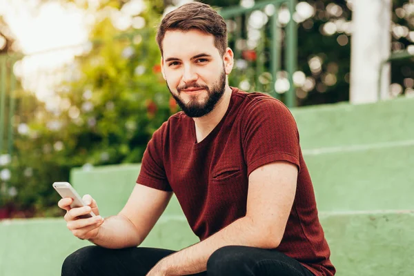 Привлекательный молодой улыбающийся мужчина пользуется телефоном в общественном парке — стоковое фото