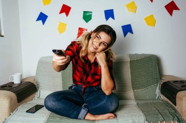 Brezilya Festa Junina 'sı evde. Tipik kıyafetler giyen biri. Renkli bayraklarla süslenmiş oturma odası. TV izleyen bir kadın..