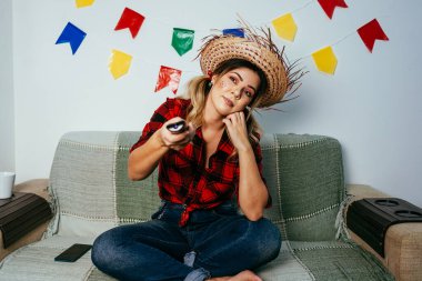 Brezilya Festa Junina 'sı evde. Tipik kıyafetler giyen biri. Renkli bayraklarla süslenmiş oturma odası. Üzgün kadın TV izliyor..