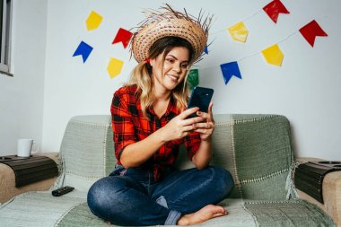 Brezilya Festa Junina 'sı evde. Tipik kıyafetler giyen biri. Renkli bayraklarla süslenmiş oturma odası. Cep telefonu kullanan kadın.