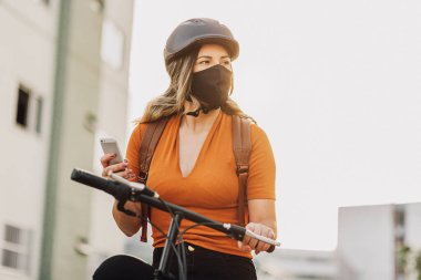 Şehirde akıllı telefon kullanarak bisiklet üstünde duran genç bir yetişkin COVID-19 'a karşı maske takıyor.