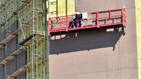 High-altitude work on the external walls of glass wool insulation. Workers use special equipment to insulate the facade of the building and finish it with panels