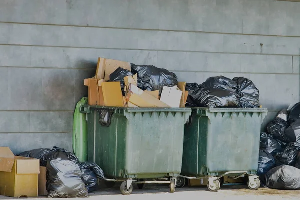 containers for garbage are overflowed with  black plastic bags with rubbish and cardboard boxes