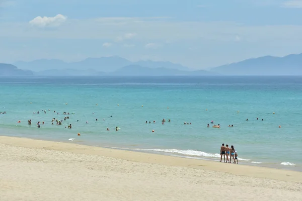 Hermosa playa de arena blanca limpia europea con agua azul sobre el fondo del cielo nublado y las montañas. Familia de cuatro personas caminando por la costa — Foto de Stock