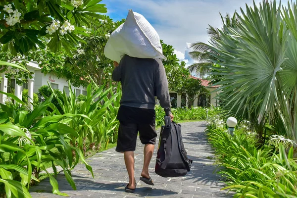 一个穿灰色毛衣和黑色短裤的男人肩上扛着一个白色的包，手里拿着一个背包。他沿着一条石头小路走在酒店周围的棕榈树和绿色植物之间。 免版税图库照片