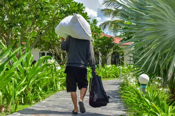 Um homem de camisola cinza e calções pretos carrega um saco branco no ombro e segura uma mochila na mão. Ele caminha ao longo de um caminho de pedra entre palmeiras e plantas verdes ao redor do hotel — Fotografia de Stock