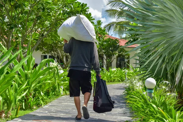 一个穿灰色毛衣和黑色短裤的男人肩上扛着一个白色的包，手里拿着一个背包。他沿着一条石头小路走在酒店周围的棕榈树和绿色植物之间。 免版税图库图片