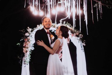 Aşık yeni evliler yavaşça kucaklamak. Düğün töreni doğada. Işıklar elektrik Garland düğün aydınlatmak.