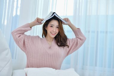 Eğitim kış konsepti. Şirin Asyalı kız kış elbise. Güzel bir kadın beyaz bir yatak odasında rahatlatıcı bir şey. Kış giyim güzel kadın yatak odasında çalışıyorlar. Yatak odasında kadın giyiyor kazak.