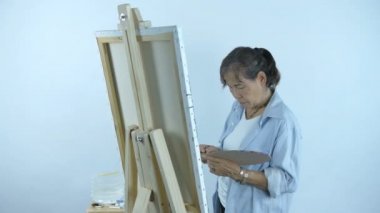 Sanatçı Kavramı. Orta yaşlı bir kadın beyaz arka planda bir tabakta renkleri karıştırıyor. 4k Çözünürlüğü.