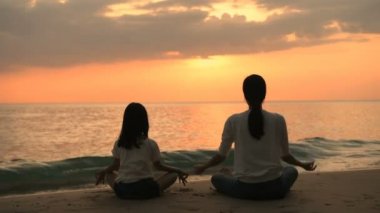Sağlık kavramları. Anne ve kızının silueti akşam deniz kenarında yoga yapıyor. 4k Çözünürlüğü.