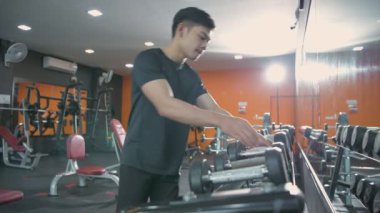 Fitness konsepti. Genç bir Asyalı adam içeri girer ve bir dambılı kaldırır. 4k Çözünürlüğü.
