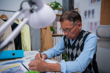 Süveter giyen yaşlı bir adam özenle çalışma alanında tasarım çiziyor..