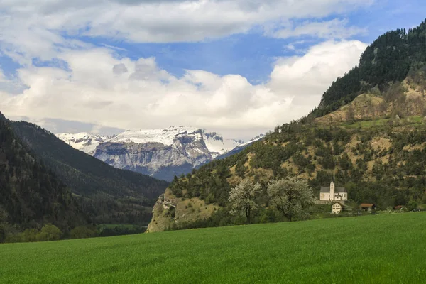 Small church at the foot of the mountain in spring. In the foreground is green meadow with a pair of flowering fruit trees. High mountains are still partly covered with snow.