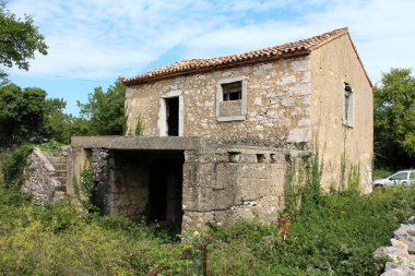 Açık teras ile kaplı ön büyük Bodrum ile yok pencere ve kapılar ile terkedilmiş geleneksel taş ev ve bulutlu mavi gökyüzü arka planda yüksek çim ve diğer bitkiler ile çevrili