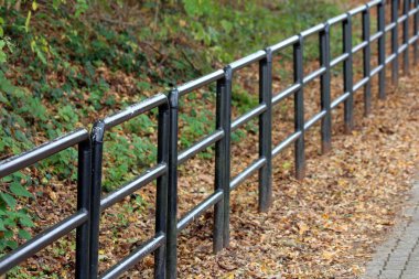 Sıcak güneşli bir gün yeşil ve kahverengi düşmüş yaprakları ile çevrili yürüyüş yolu yanında güçlü borulardan yapılmış siyah metal koruyucu çit