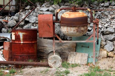 Sıcak güneşli bir gün yerel arka bahçede taşlar ve çöp ile çevrili tamamen paslanmış metal varil ve inşaat malzemesi yanında paslı Vintage elektrik beton mikser
