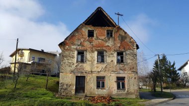 Kırık pencereler, duvarlar düşmüş cephe ve tuğla, asfalt yolun yanında ve arka planda modern banliyö evi ile terkedilmiş eski harap ev