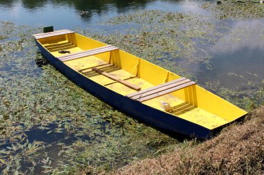 Nehir kıyısına bağlı oturma ve sıcak yaz gününde düşmüş yaprakları ve nehir bitki örtüsü ile çevrili olarak üzerinde panoları ile mavi ve sarı ahşap nehir teknesi