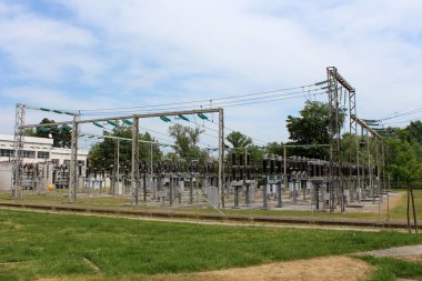 Metal çit ve sıcak güneşli bir gün arka planda ağaçlar ve bulutlu mavi gökyüzü ile yüksek kesilmemiş çim ile çevrili yoğun teller için cam ve seramik izolatörler ile bağlı birden fazla elektrik enerjisi transformatörleri ile endüstriyel güç santrali
